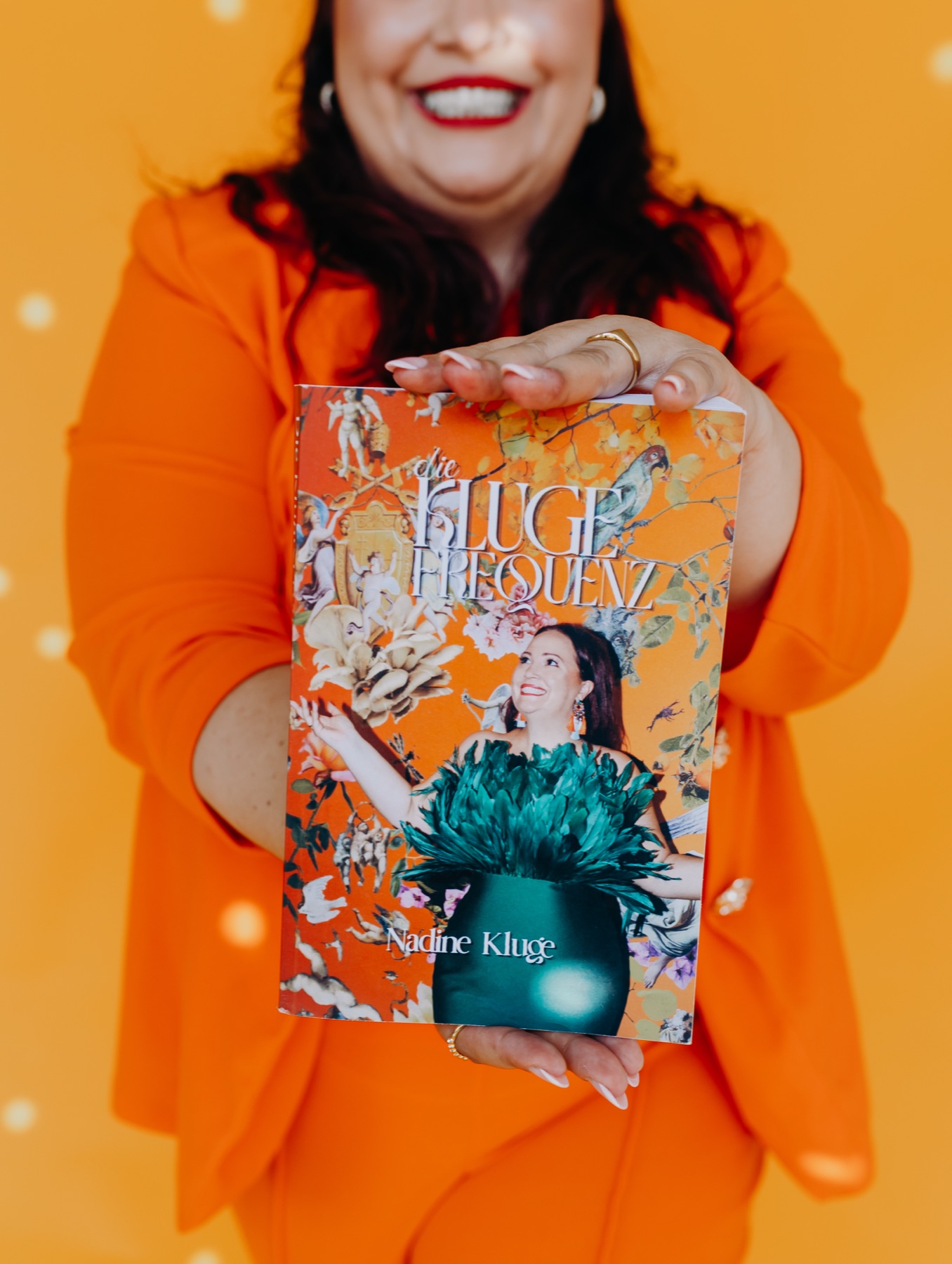 Nadine Kluge im orangen Anzug hält ihr Buch „Die Kluge Frequenz“ in die Kamera